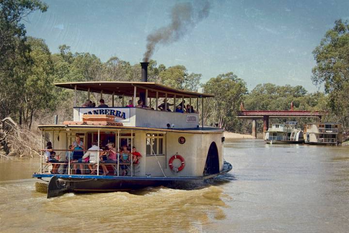The PS Canberra paddle steamer carries tourists on a brown river, with eucalyptus trees lining the banks and other paddle steamers moored at a dock in the background.