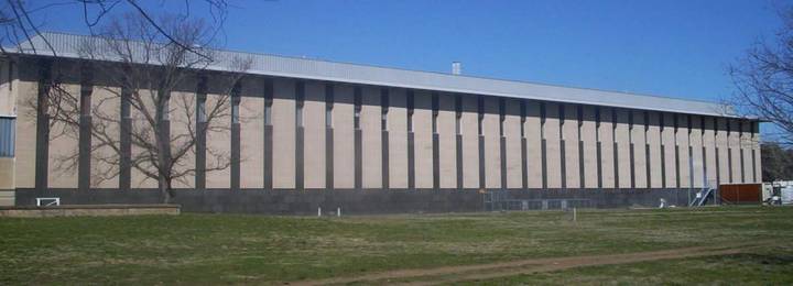 The exterior of the Royal Australian Mint, a long low-rise beige building with regularly spaced dark vertical pillar elements and a metal roof, photographed from across a grass law