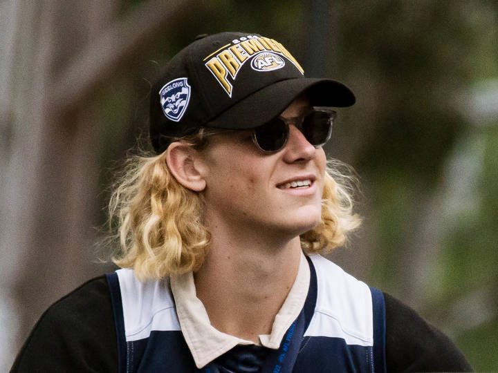 A young man with long blonde wavy hair wearing a black AFL Premiers cap and sunglasses, smiling and glancing upward, dressed in a navy and white sports top.