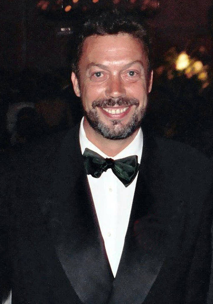 A man with dark curly hair and a short beard wearing a black tuxedo and dark green bow tie smiles broadly at the camera at what appears to be a formal evening event.