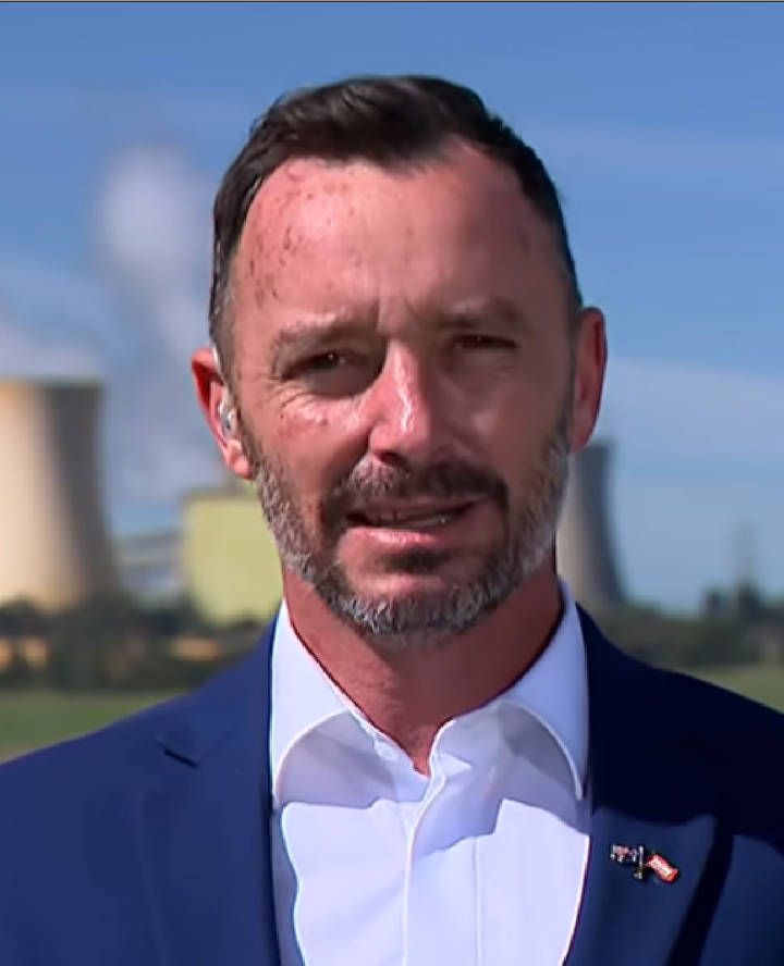 Warren Pickering. A man in a blue suit and white shirt with a flag lapel pin stands outdoors with industrial cooling towers visible in the background under a blue sky.
