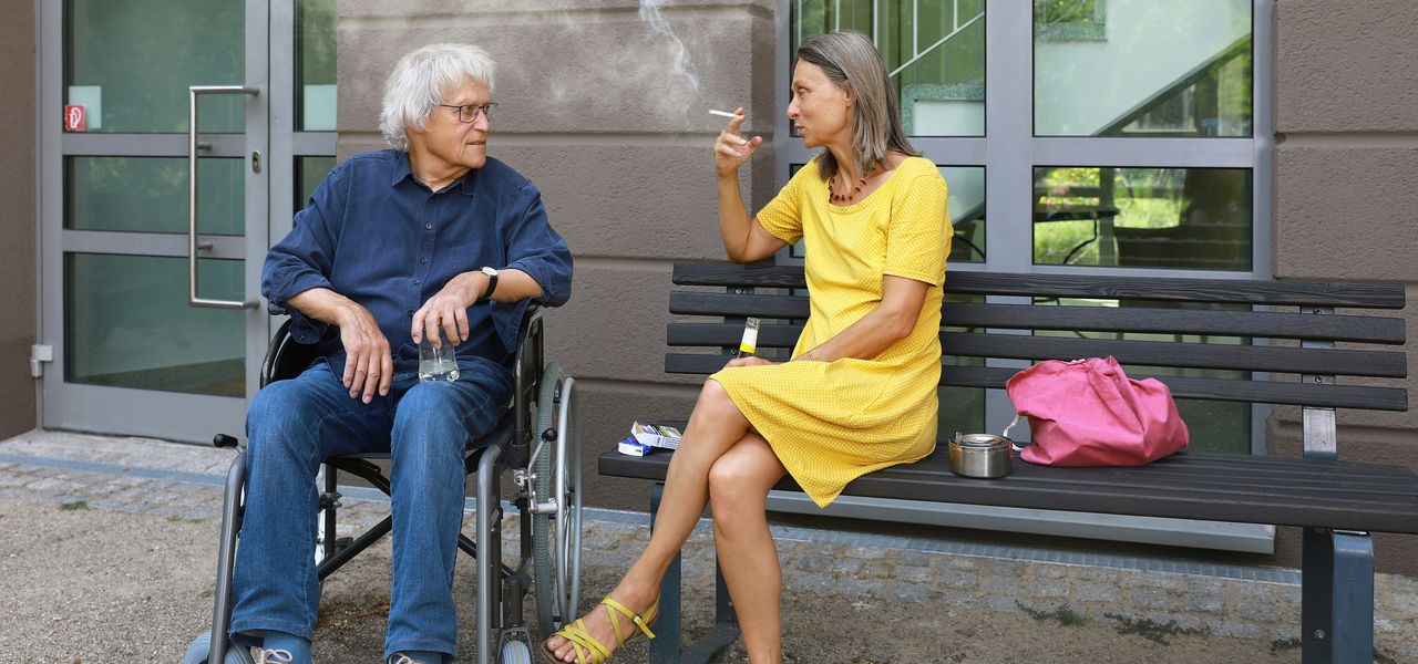 a man sitting on a bench next to a woman smoking a cigarette