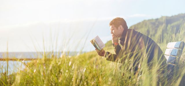 man reading book on beach near lake during daytime