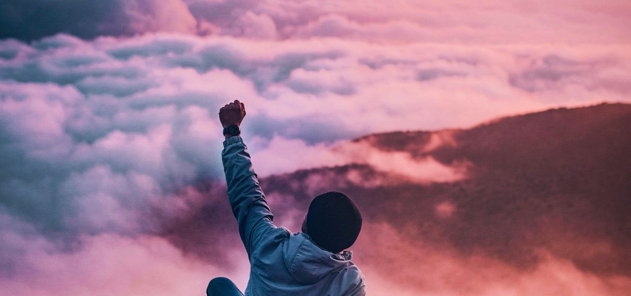 man sitting on mountain cliff facing white clouds rising one hand at golden hour