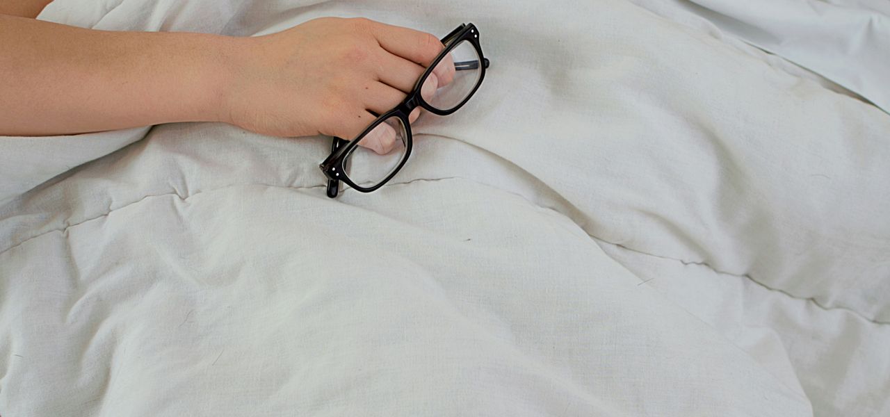 person lying on bed while covering face with pillow and holding eyeglasses