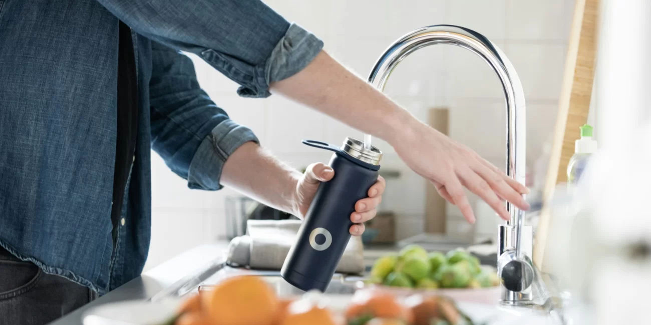 Person filling a water bottle from a kitchen faucet, symbolizing fluoride exposure through tap water. Fresh fruits and vegetables are seen on the counter in the background.