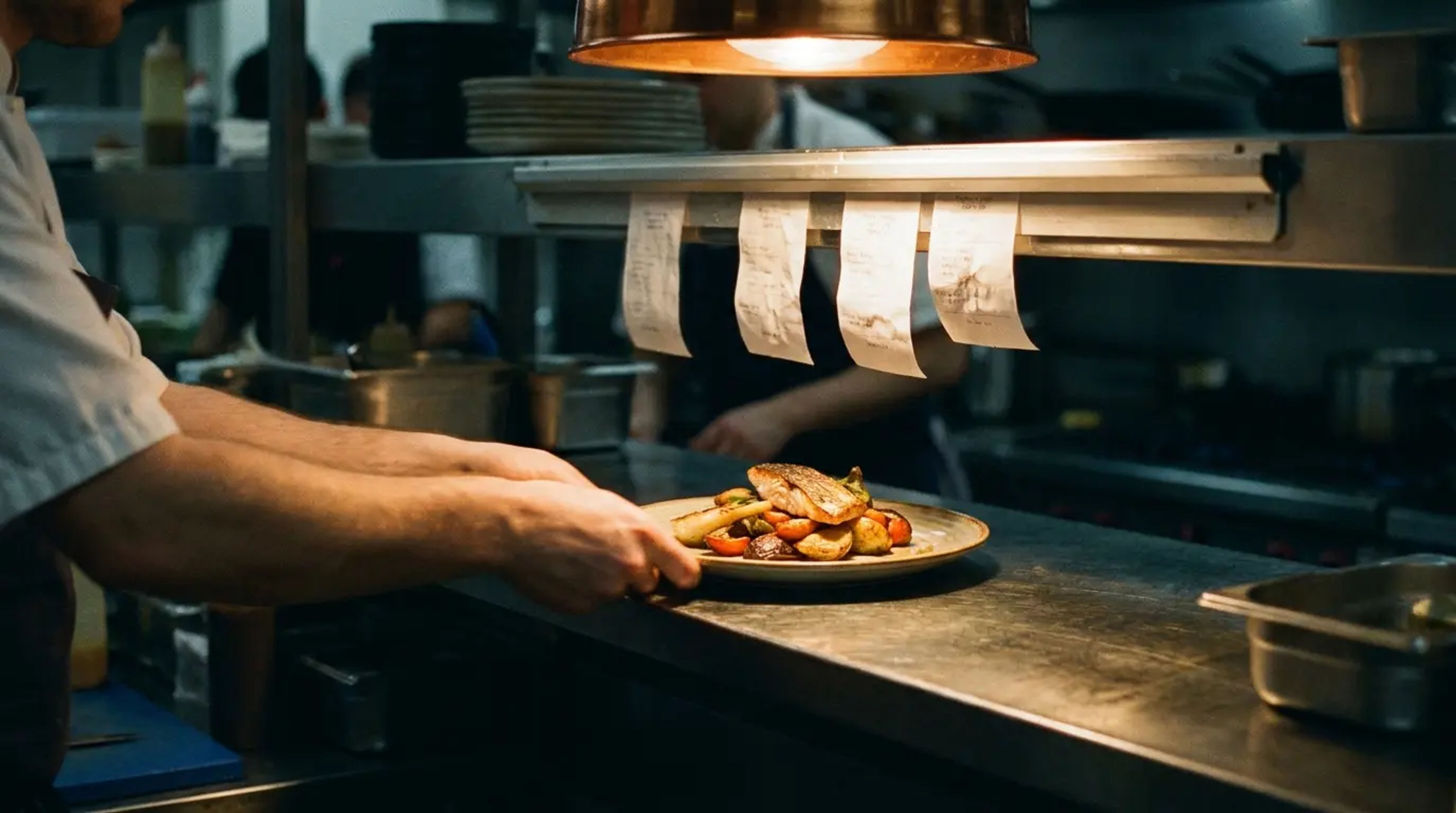A chef reaching a plated dish onto the pass beneath a row of order tickets