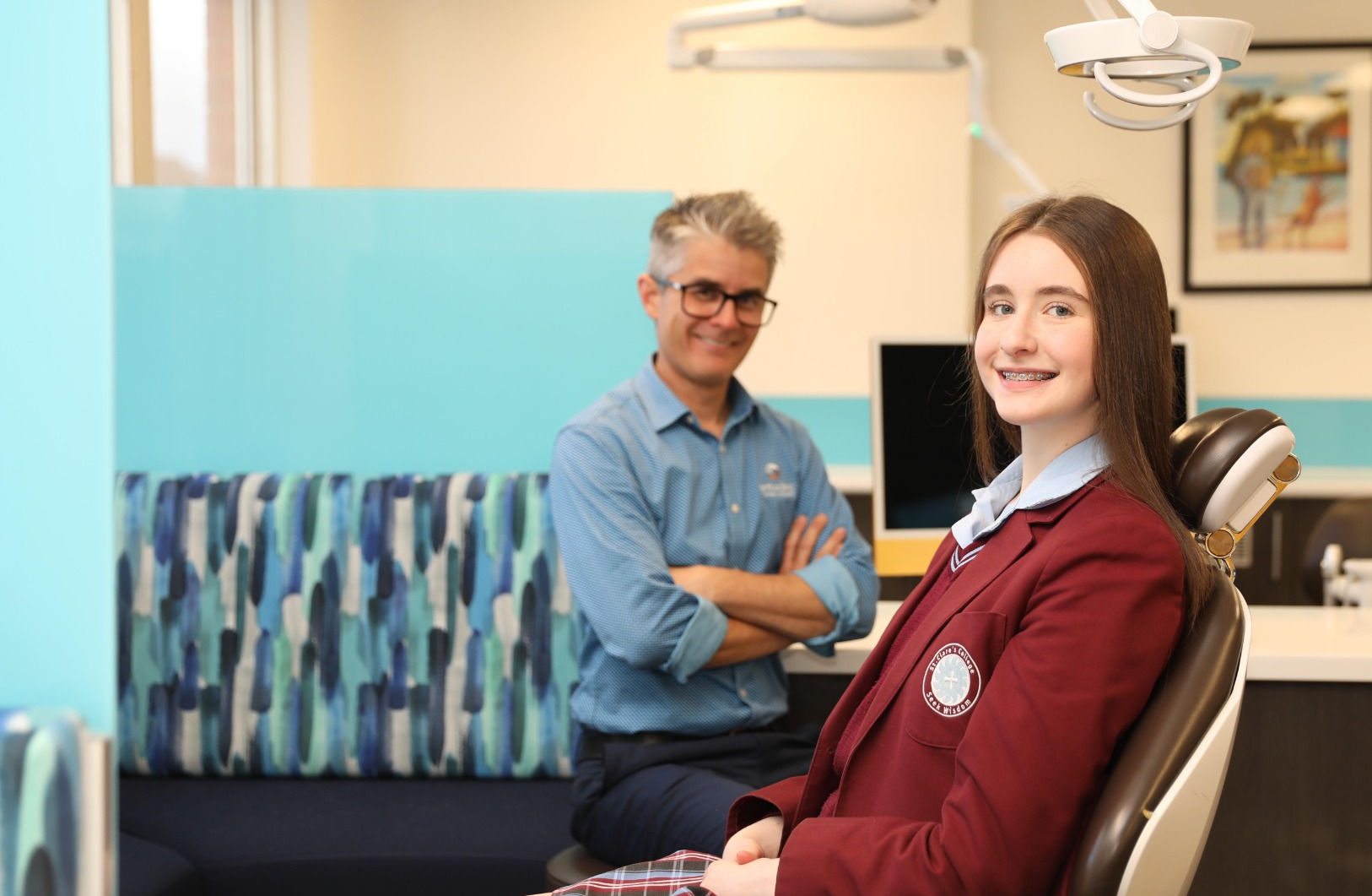 Teenage girl in school uniform sitting in dentist chair smiling at camera.  Smiling dentist in the background
