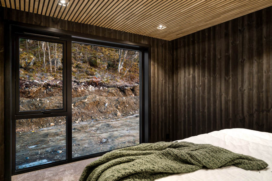 Moderne soverom med mørke trevegger, lyssatt spilehimling og en stor glassdør med utsikt mot naturen. En grønn strikket pledd ligger på en seng med hvitt sengetøy, som skaper en lun atmosfære