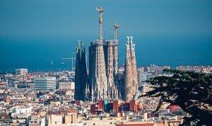 Barcelona panorámája a magasból, a háztetők közül kiemelkedő, még építés alatt álló Sagrada Família bazilikával és darukkal, a háttérben a tengerrel, tiszta kék égbolt alatt.
