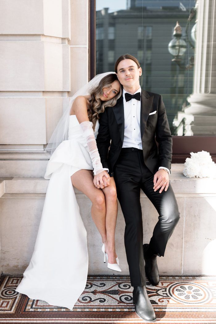A couple in wedding attire sit against a building holding hands.
