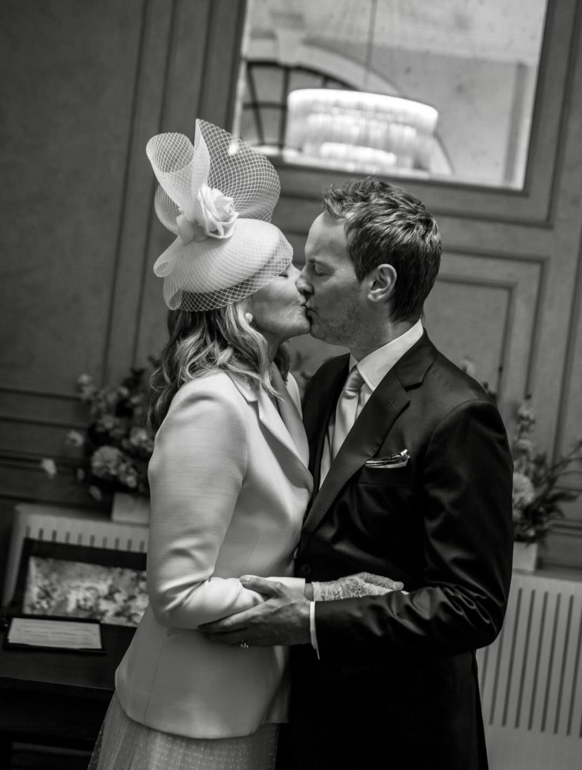 Kim Cattrall kissing her husband at their wedding wearing a white hat and blazer dress