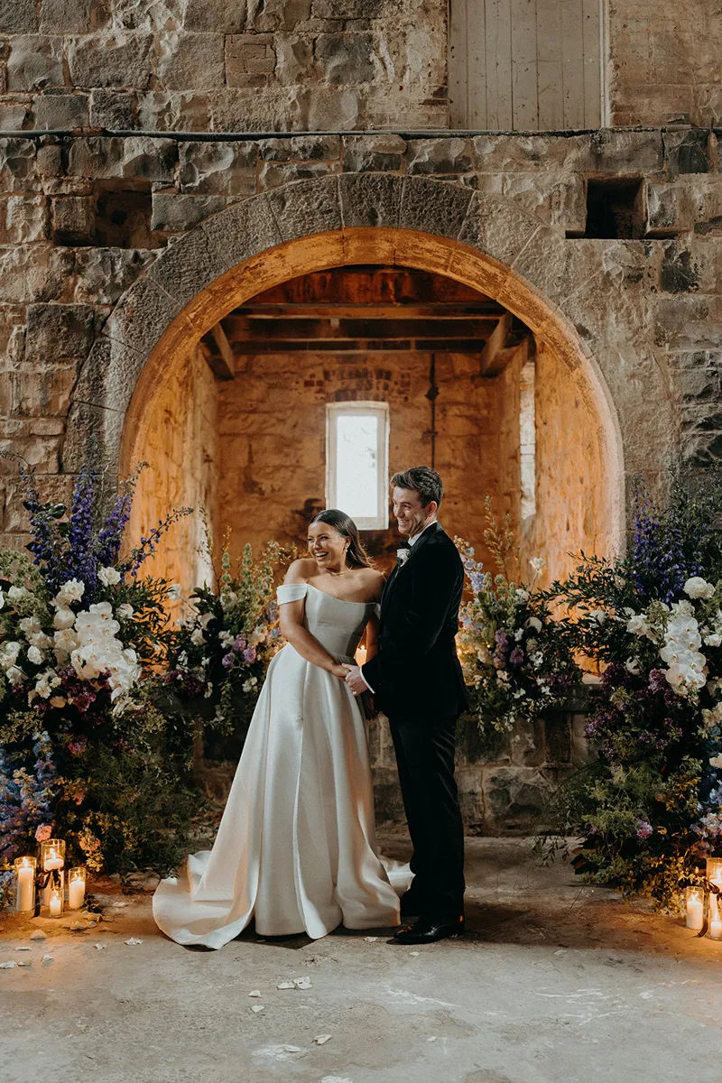 A couple smiling in front of a lavender floral arrangement during their wedding ceremony.