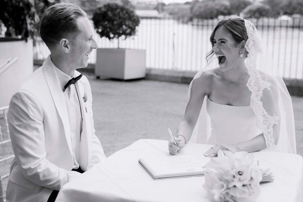 A black and white photo of a bride open-mouth smiling at her groom while signing wedding paperwork.