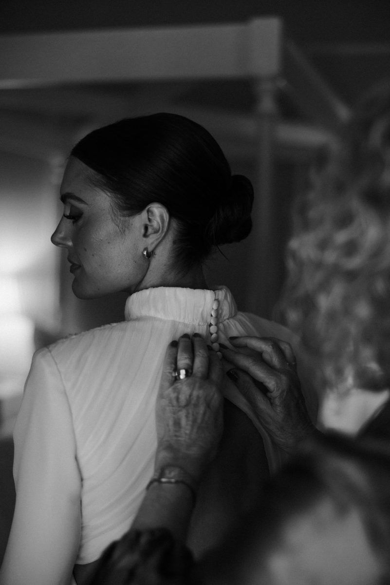 Elderly hands doing up the back of a bridal gown, while the bride looks over her shoulder.