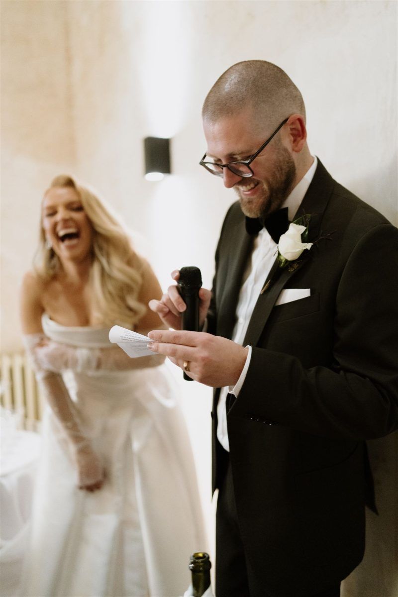 A close up of a groom reading a speech, holding a microphone and laughing, with his bride laughing out of focus behind him.