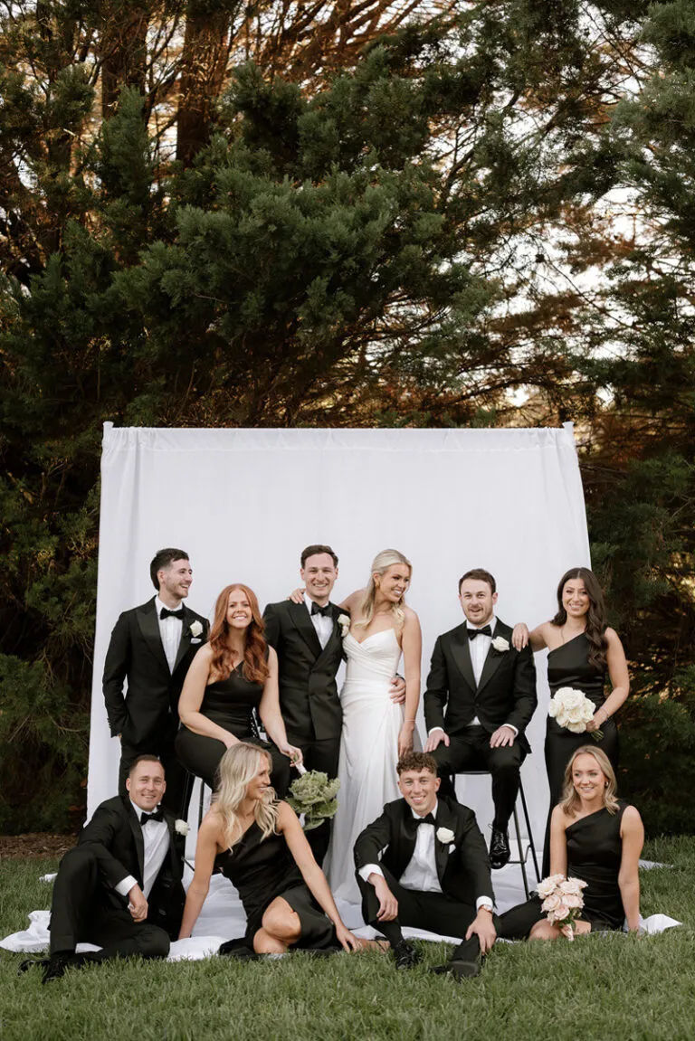 A bride and groom are surrounded by their bridal party, all in black, against a white sheet backdrop.