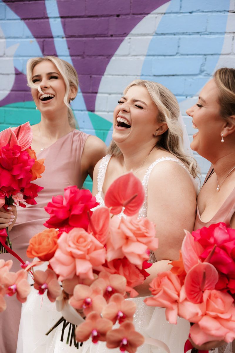 Close up of a bride and two bridesmaids holding bright pink, fushia and peach bouquets against a brightly painted brick wall.