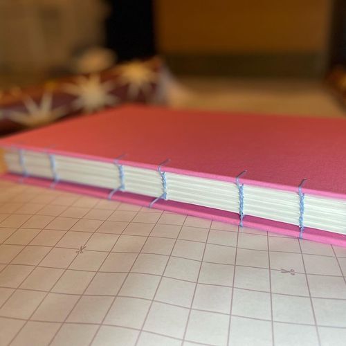 A pink handmade sketchbook, bound with coptic stitch. The cover is a light pink, and the stitches are in a light blue.