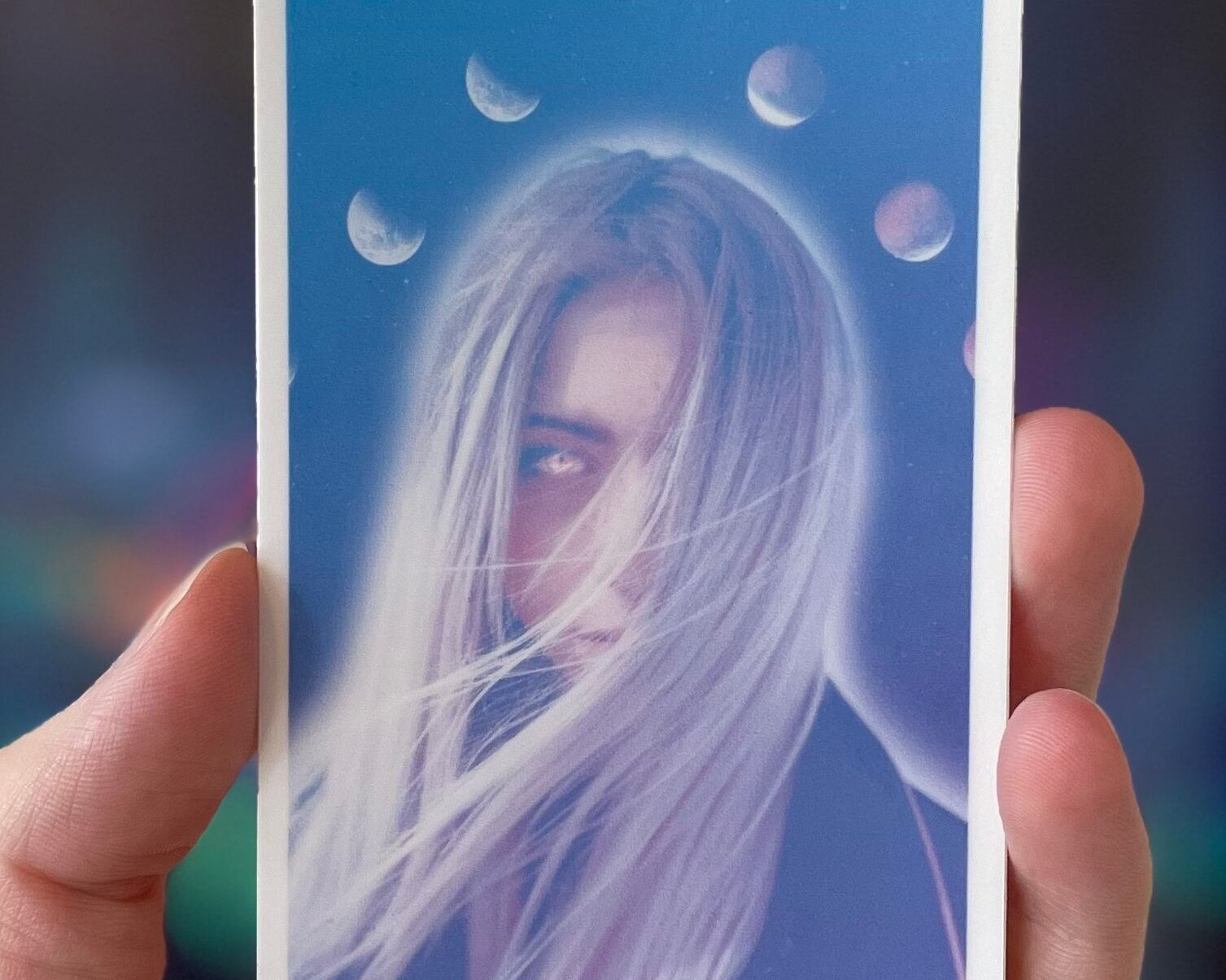 My hand, holding up a tarot-sized tarot sticker. It is the High Priestess, and shows a woman with long hair that is being messed with by wind. She is glowing, and her one visible eye is golden. Above her are the phases of the moon.