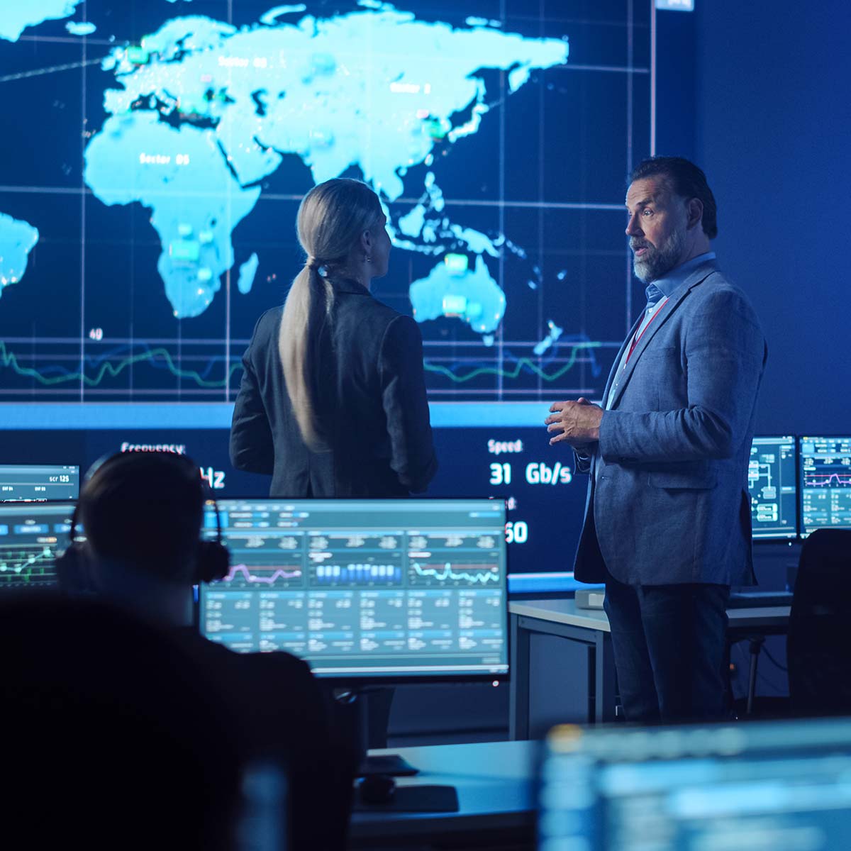 man in control room explaining to new woman hire maps in background