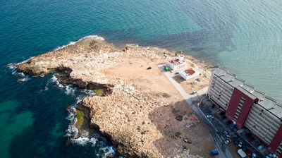 Coastal Majesty in Torrevieja: Witness the harmonious dance of nature's power and human resilience in this captivating aerial view of a rugged cliff, majestic lighthouse, and bustling seaside town.