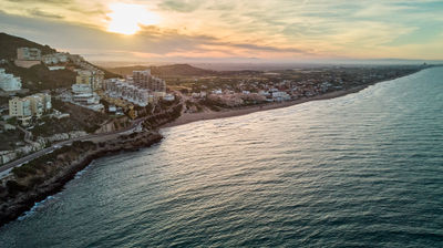 Sunset beach view: long sandy stretch with buildings along coast. Skyline in top left, cliffs in bottom left, lone boat in bottom right. Serene scene.