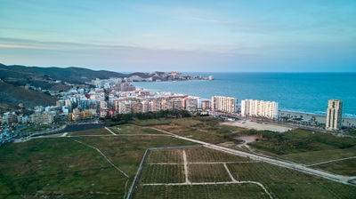 Urban metropolis near ocean with skyscrapers, bustling streets and cemetery; hilltop offers panoramic view of city and coastline; sandy beach to south.