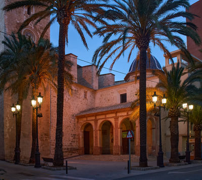 The image depicts an urban street scene at dusk, with tall palm trees lining both sides of the road and a church in the centre