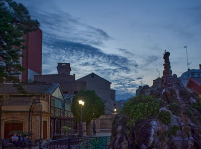 Cityscape charm at dusk: A majestic fountain takes center stage as people stroll through a vibrant city, embraced by towering clock towers and painted by a serene dusky sky.