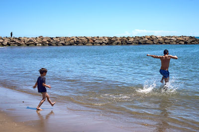 A man and a young boy enjoy a sunny day on a sandy beach. The boy follows his father running into shallow water. Joyful scene of the summer play.