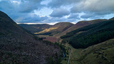 An aerial view of Glen Doll - mountains, pine forest on slopes and a road under evening sky