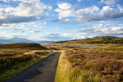 A road, rich fields, and trees may be seen in Scotland's Glen Quaich beneath a picturesque sky.