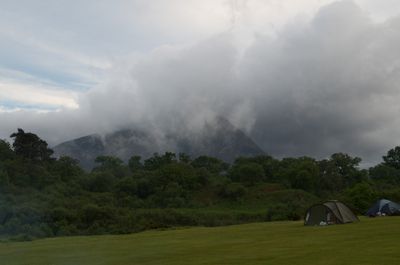 Isle of Arran, Scotland. An idyllic field, surrounded by mountains, with a tent in the center. A person stands nearby, trees fill the landscape, creating a peaceful and solitary connection to nature.