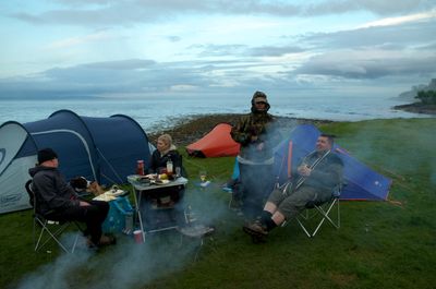 Isle of Arran, Scotland. A group of people relax on a beach, enjoying a camping trip. They sit under a tent, accompanied by dogs, while food cooks on a grill nearby.