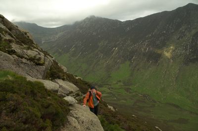 Isle of Arran, Scotland. A man in an orange jacket stands on a mountain summit, surrounded by foggy and ethereal scenery. He walks along a rugged path, appreciating the beauty of nature.