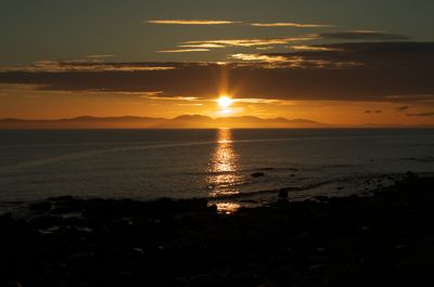 Kintyre, Scotland. A majestic sunset over water with a mountain in the distance. Clouds add depth to the vibrant sky, creating a captivating and picturesque scene.