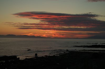 Kintyre, Scotland. A picturesque sunset scene with a person standing on a rocky beach, silhouetted against a vibrant sky. Serene and beautiful, capturing the colors where the sky meets the water.