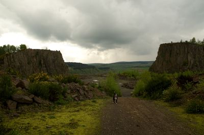 A serene country landscape with a dirt road, cloudy sky, person blending in, sign, rock cliff, trees, dog, green bushes, rocky terrain. Tranquil rural beauty.