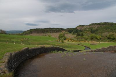 A serene landscape with a river, stone wall, grassy field, trees, hills, and cloudy sky. Beauty and tranquility of nature.