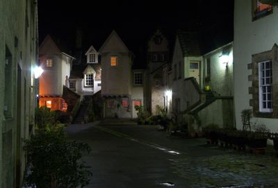 A peaceful, quaint neighborhood at night is depicted by a wet street, illuminated by streetlights, with houses, a tower, and a solitary tree.