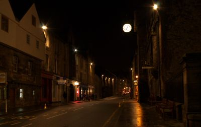 A vibrant city street at night with illuminated buildings, streetlights, and a prominent clock. Nighttime ambience blends activity and calmness.