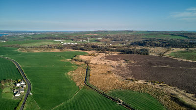 Aerial view of distant green and brown fields, with the sea in the distance. A few houses in the cou