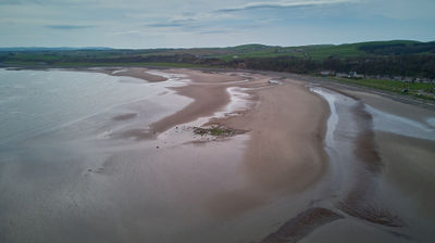 Aerial image of the sea and a sandy beach against a blue sky. Green fields and hills may be seen fro