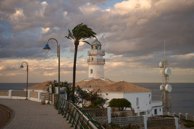 a lighthouse on a cliff overlooking the ocean with a cloudy sky and a palm tree in the foreground