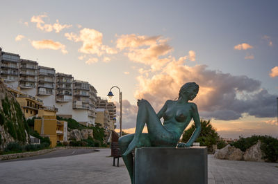 Cullera, Valencia, Spain - 03.13.2023 a statue of a woman sits on a bench in front of a building with a cloudy sky in the background