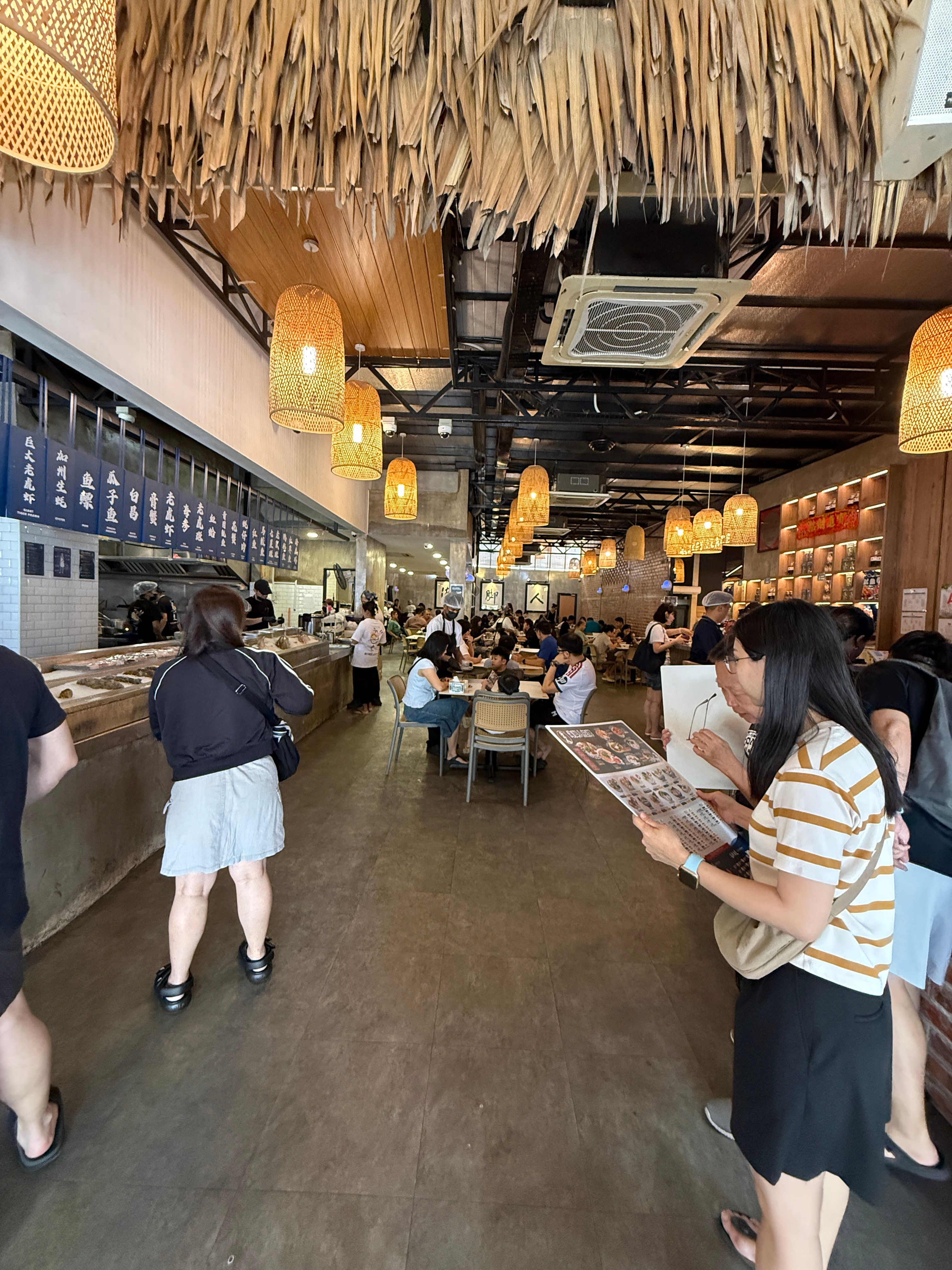 Busy Hai Kah Lang interior — thatched ceiling, woven pendant lights, diners and staff along the seafood counter