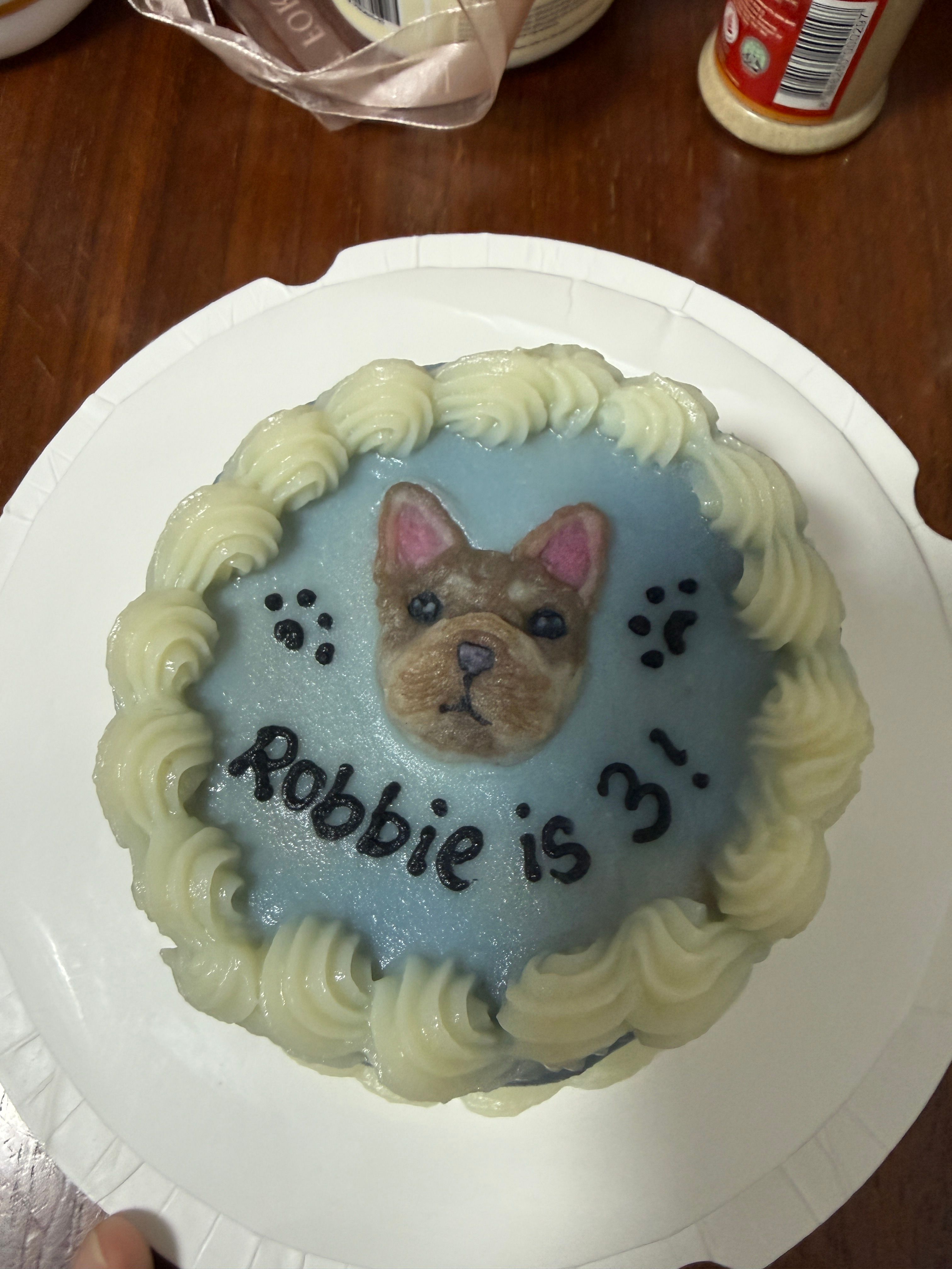 Overhead view of Robbie's light blue dog birthday cake with white piped border, French Bulldog portrait, paw prints, and Robbie is 3 icing