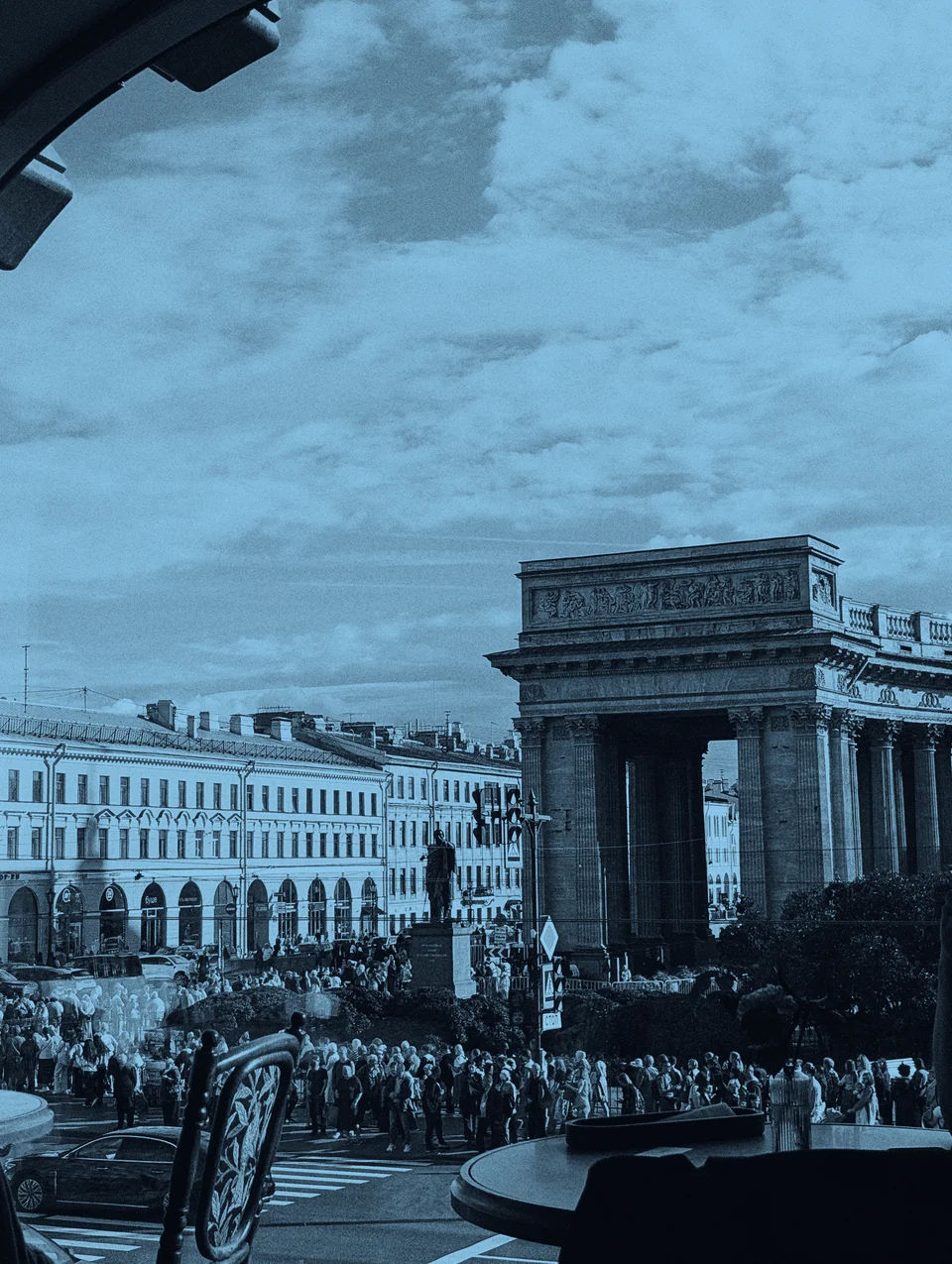 View from a cafe window, with tables and chairs in the foreground overlooking a crowded street and a monumental stone colonnade under a cloudy sky.