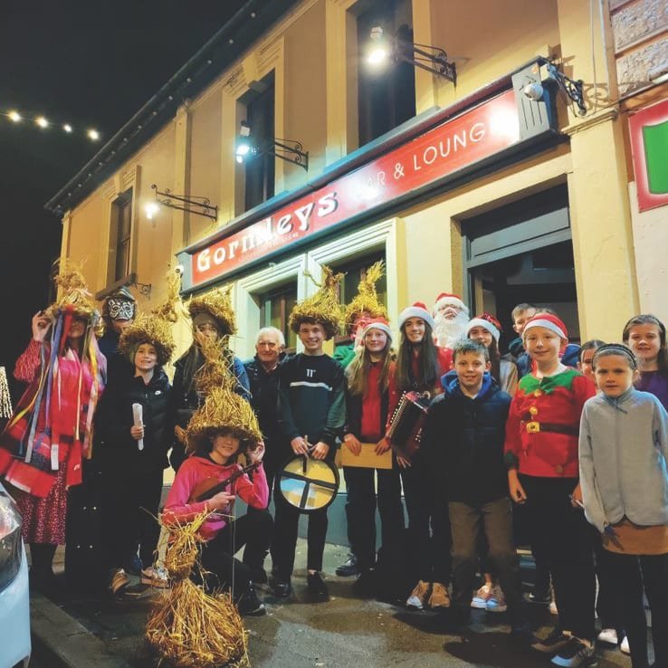 Outside Gormleys Bar and Lounge a large group of children and young adults wearing costumes including Santa hats, hats covered in straw, a mask and a white Santa beard, a few are holding instruments